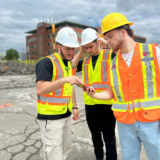 Three of Brixl's co-founders in hard hats and safety vests on a construction site, looking at a phone together.